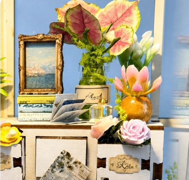Sur le meuble clair, le cadre doré côtoie des livres, un petit vase et des feuillages colorés. Les fleurs se répartissent à différentes hauteurs pour animer ce coin de la serre.