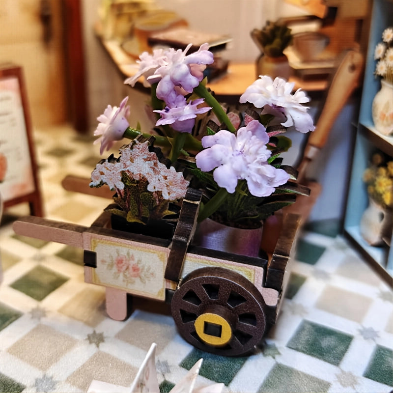 Le chariot rassemble plusieurs pots de fleurs mauves et claires. Placé au rez-de-chaussée, il donne à la scène l’allure d’une composition en cours.